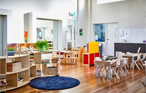 View of a modern playroom at a daycare with the tables and chairs on the right side of the room and shelving with various items and baskets on it along with a small round blue rug on the left side.