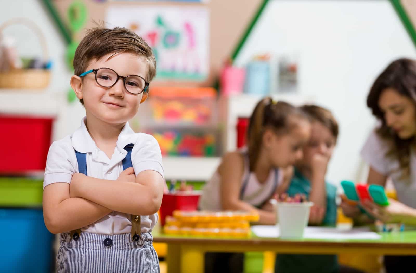 A little boy with suspenders and glasses crossing his arms smiling with his peers blurred, playing in the background.