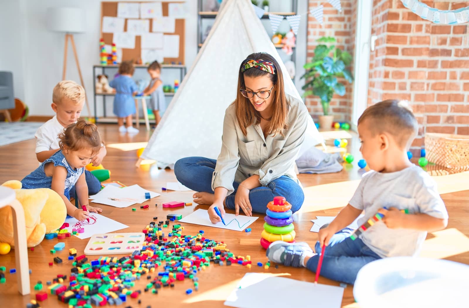 Teacher at a daycare sitting on the ground cross legged and drawing on pieces of paper with three other toddlers as they sit around a pile of lego bricks.